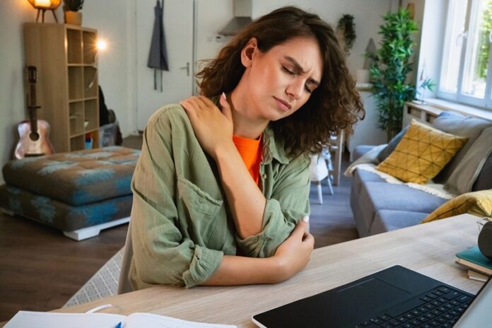 Young Caucasian woman, having a pain in her shoulder, while e-learning at home