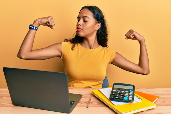 rethinking employee benefits: a professionally-dressed business woman flexing her biceps while sitting at her laptop.