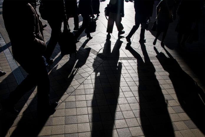 A photo of silhouettes of commuters heading to work from a train station. The morning light makes them cast long shadows.